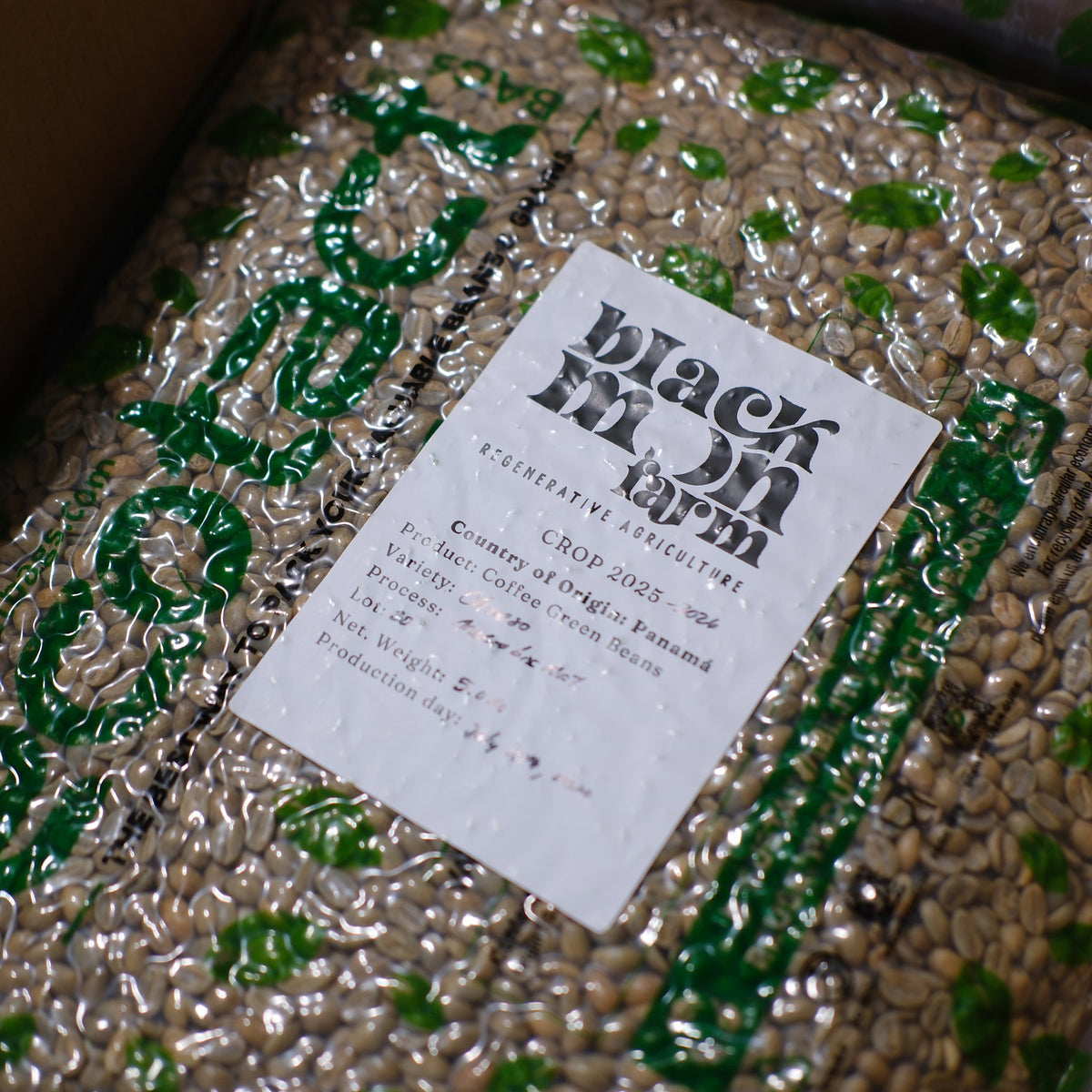 A close-up of packaged coffee beans with a white label reading 'Black Moon Farm' and 'Cup of Coffee from Panama,' surrounded by green and silver reflective wrapping.