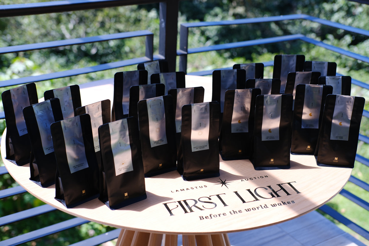 Multiple black coffee bag packages with clear front windows are arranged on a round wooden table outdoors. The table is branded FIRST LIGHT with smaller text including ANASTASIO AUSTIN.