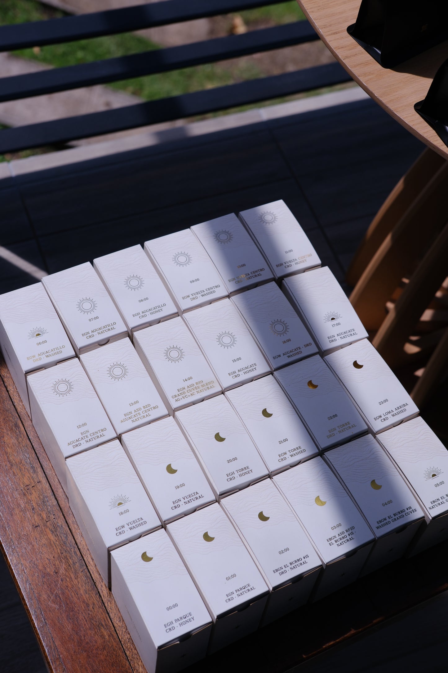 A stack of small white product boxes arranged on a wooden table outdoors. Each box has a small printed logo and text, with one box showing a gold crescent moon symbol.