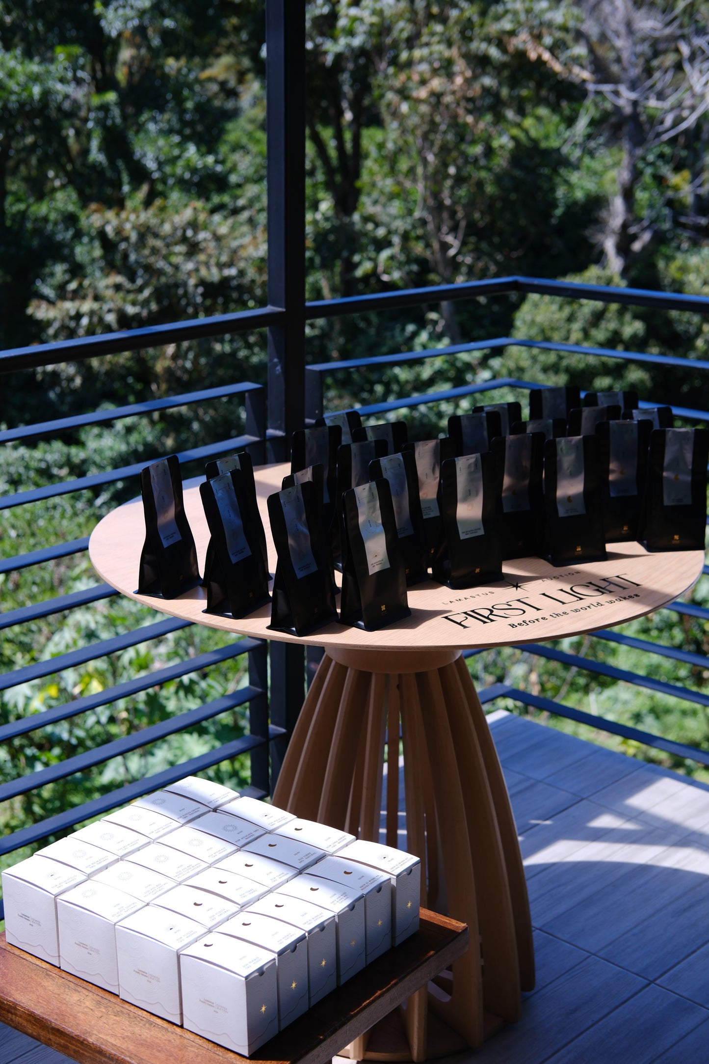 A display of black rectangular phone cases on a round wooden table, with a stack of white product boxes on a wooden bench in the foreground. The setting is outdoors on a balcony with a railing and a lush green forest background.