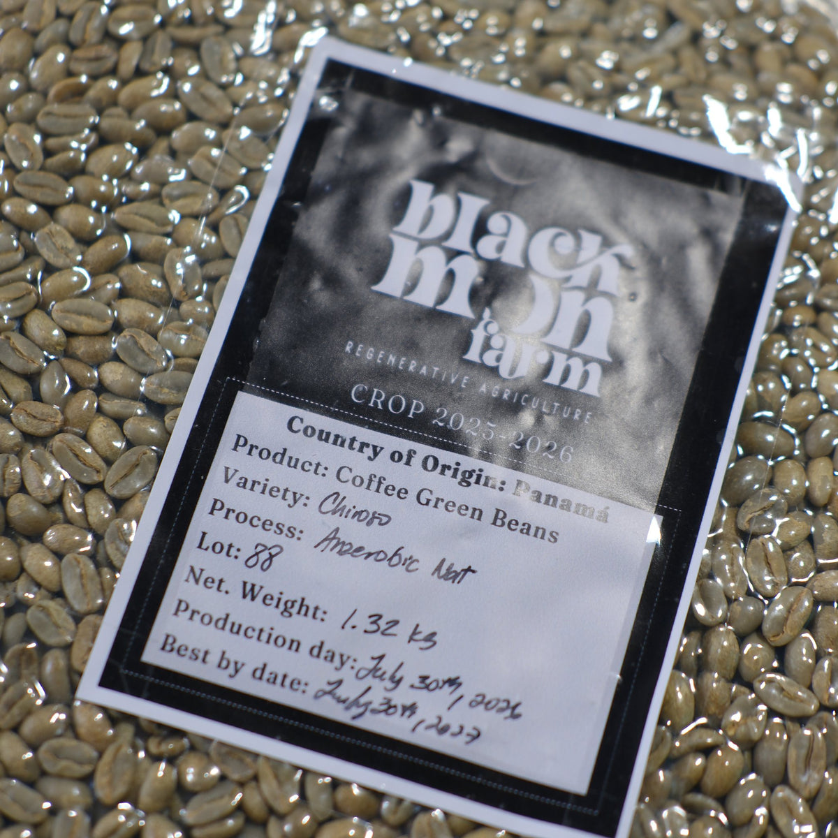 A close-up of a product label for Black Mdn Farm Coffee Green Beans, placed on top of loose green coffee beans. The label details the crop year, variety, and other production information.