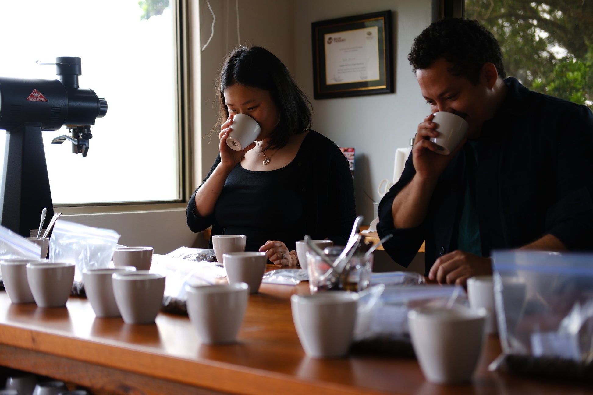 A man and a woman sit at a wooden table in a bright room, each holding a white ceramic mug. Several white mugs and papers are arranged on the table, with a coffee roaster visible in the background near a window.